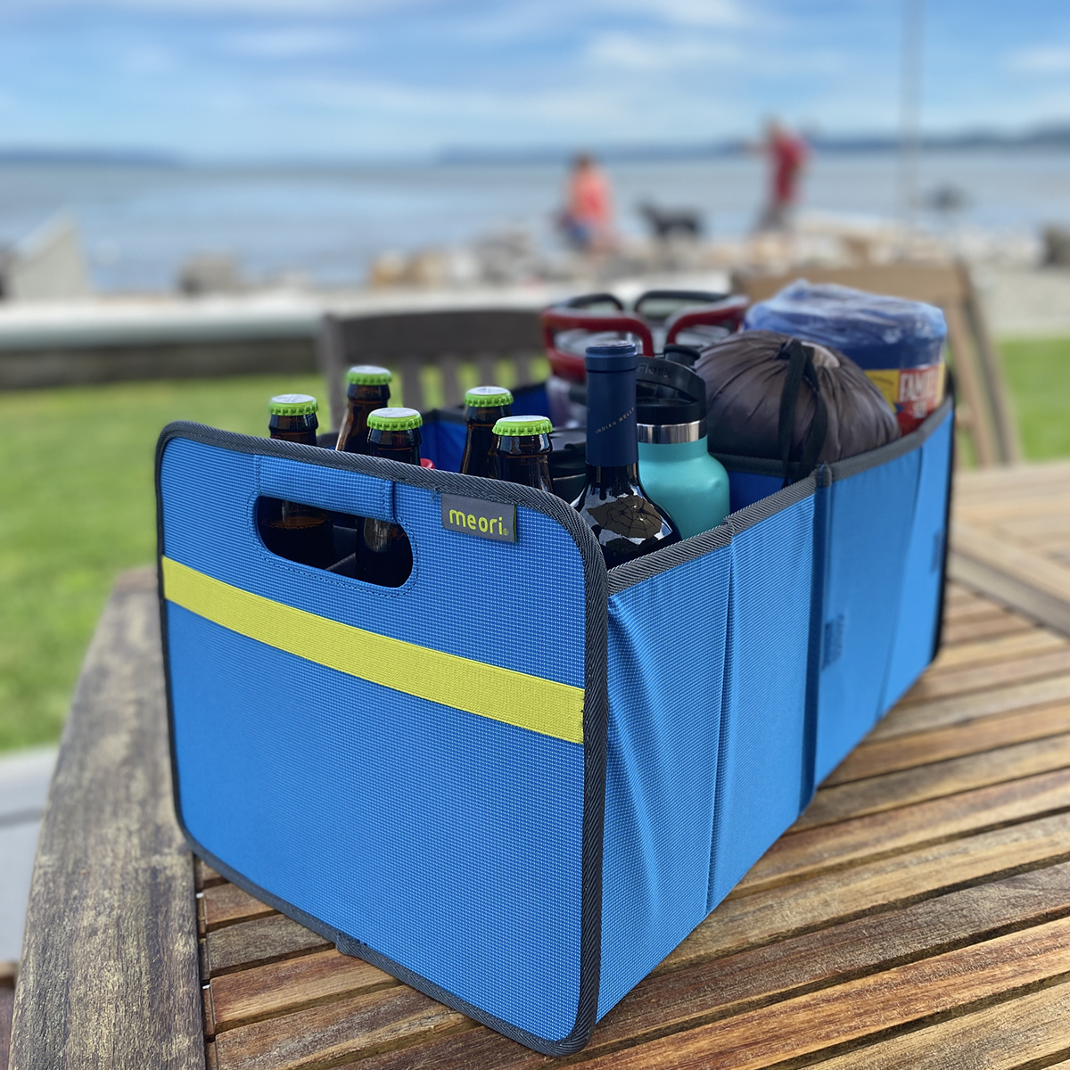 B100844-10-meori-Premium-Box-with-Bottle-Insert-Med-Blue-LST Blue Car Trunk Organizer with 9 bottles and camping gear on a teak table with a beach in the background