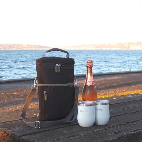 Black Wine Gift Bag next to champagne bottle and two insulated wine tumblers on picknic table by the beach